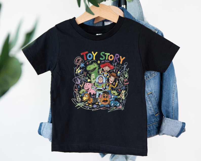 May include: A black short-sleeved t-shirt featuring a colorful graphic print of Toy Story characters including Woody, Buzz Lightyear, Jessie, Rex, Hamm, and Mr. Potato Head, surrounded by scribbled line art and the text Toy Story and yee haw.