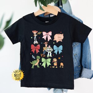 May include: Black t-shirt with a graphic print of Toy Story characters wearing bows. The characters include Woody, Buzz Lightyear, Rex, and Hamm. The text "Ships Next Day" is visible in the lower left corner of the image.