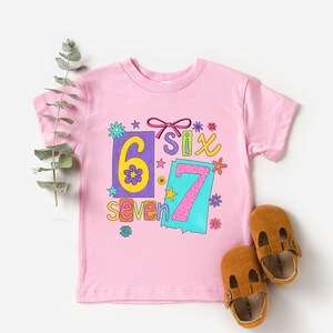 May include: A pink t-shirt with the numbers "6" and "7" in colorful, playful designs. The word "six" is also displayed. Brown leather baby shoes are next to the shirt. The shirt is decorated with flowers and stars.