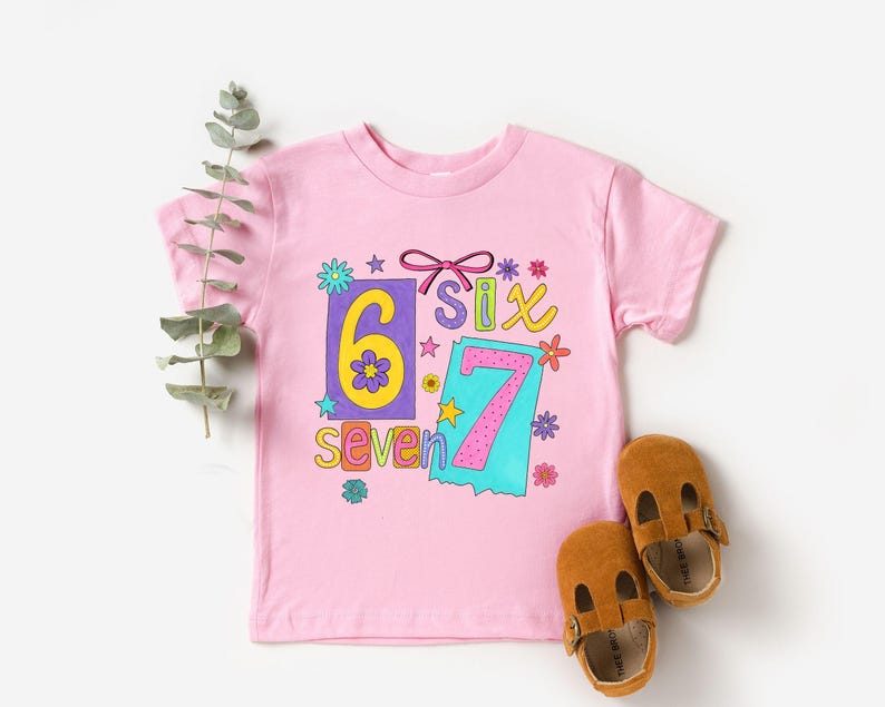 May include: A pink t-shirt with the numbers "6" and "7" in colorful, playful designs. The word "six" is also displayed. Brown leather baby shoes are next to the shirt. The shirt is decorated with flowers and stars.