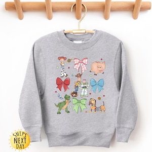 May include: Gray sweatshirt with a colorful print of Toy Story characters wearing bows. The characters include Woody, Buzz Lightyear, Jessie, Rex, and Slinky Dog.