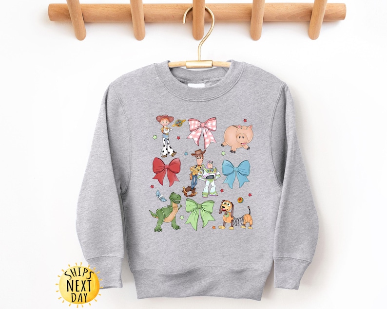 May include: Gray sweatshirt with a colorful print of Toy Story characters wearing bows. The characters include Woody, Buzz Lightyear, Jessie, Rex, and Slinky Dog.