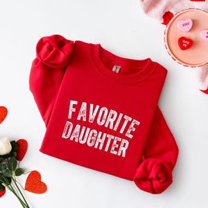 May include: A red sweatshirt with the text "FAVORITE DAUGHTER" in white, distressed lettering. The sleeves are rolled up. The image also includes heart-shaped decorations and a pink sweater with red hearts.