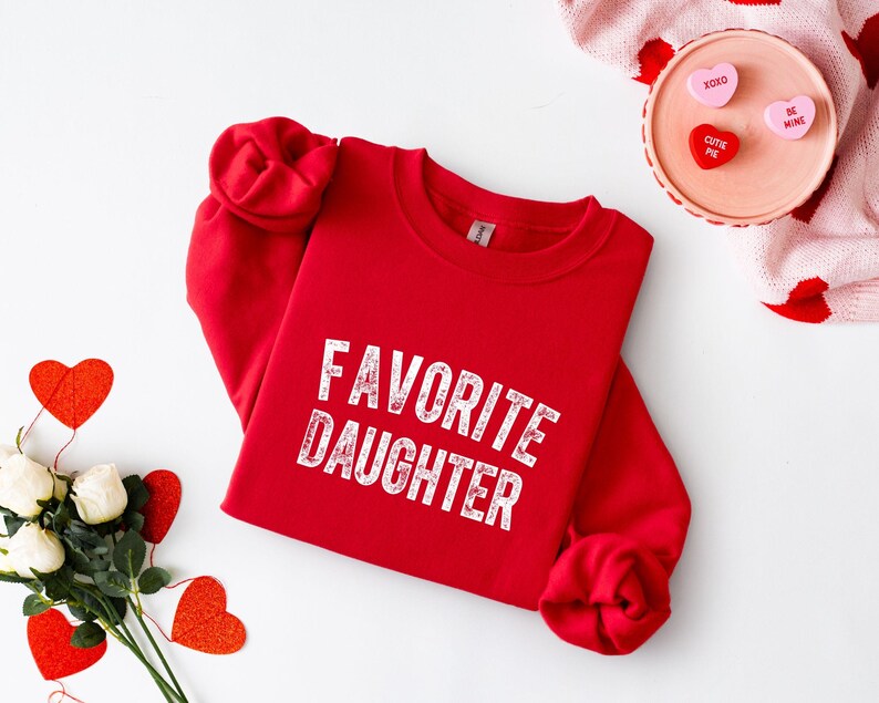 May include: A red sweatshirt with the text "FAVORITE DAUGHTER" in white, distressed lettering. The sleeves are rolled up. The image also includes heart-shaped decorations and a pink sweater with red hearts.