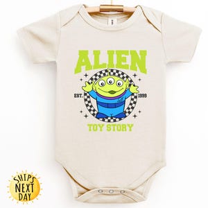 Puede incluir: Un body de beb&eacute; de color crema con un gr&aacute;fico verde, azul y negro del personaje extraterrestre de Toy Story. El gr&aacute;fico incluye el texto "Alien", "Est. 1999", y "Toy Story".