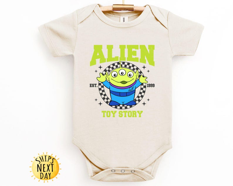 Puede incluir: Un body de beb&eacute; de color crema con un gr&aacute;fico verde, azul y negro del personaje extraterrestre de Toy Story. El gr&aacute;fico incluye el texto "Alien", "Est. 1999", y "Toy Story".