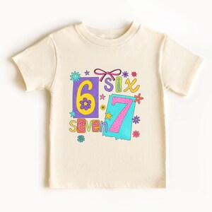 May include: Cream-colored t-shirt featuring the numbers "6" and "7" with the words "six" and "seven" in colorful, playful fonts. The design includes flowers, stars, and a bow, creating a cheerful, birthday-themed graphic.
