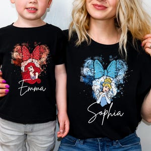 May include: Two black t-shirts featuring watercolor-style graphics of Disney princesses. The left shirt displays Ariel with a red bow and the name Emma. The right shirt displays Cinderella with a blue bow and the name Sophia in white script.