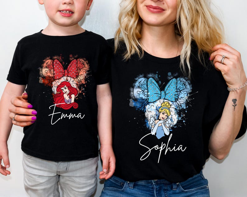 May include: Two black t-shirts featuring watercolor-style graphics of Disney princesses. The left shirt displays Ariel with a red bow and the name Emma. The right shirt displays Cinderella with a blue bow and the name Sophia in white script.