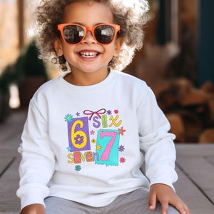 May include: White long-sleeved sweatshirt with a colorful graphic that says "6 Six 7 Seven". The design includes the numbers 6 and 7 in rectangular boxes, with flowers and stars. The sweatshirt is for children.
