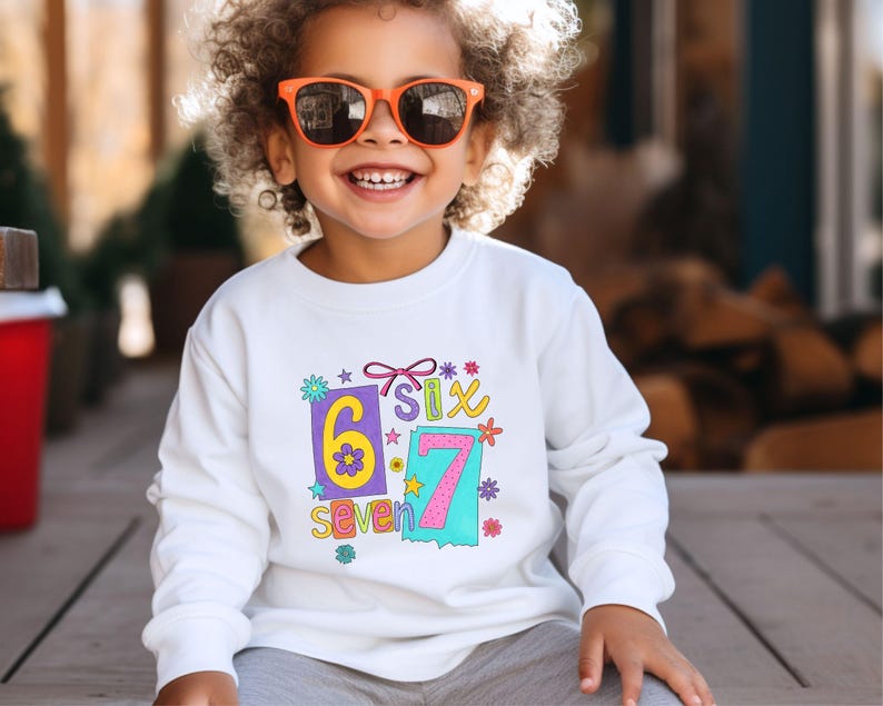 May include: White long-sleeved sweatshirt with a colorful graphic that says "6 Six 7 Seven". The design includes the numbers 6 and 7 in rectangular boxes, with flowers and stars. The sweatshirt is for children.