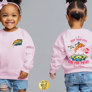 May include: A light pink sweatshirt featuring a Pizza Planet design. The front has a small graphic of the Pizza Planet aliens with a slice of pizza. The back displays the text "KEEP YOUR EYES ON THE PRIZE" with a claw machine and more aliens.