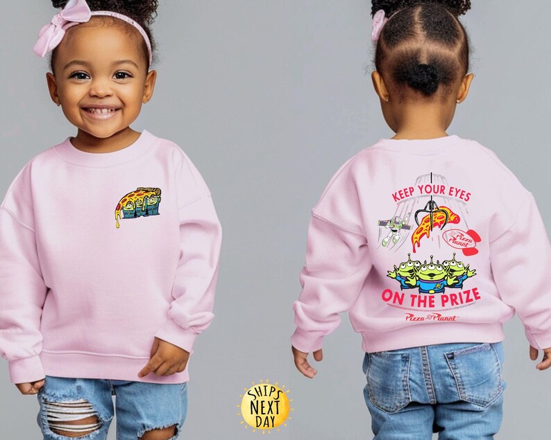 May include: A light pink sweatshirt featuring a Pizza Planet design. The front has a small graphic of the Pizza Planet aliens with a slice of pizza. The back displays the text "KEEP YOUR EYES ON THE PRIZE" with a claw machine and more aliens.