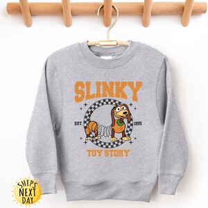 May include: Gray sweatshirt with a graphic of Slinky Dog from Toy Story. The graphic features Slinky Dog in a checkered circle with the text "Slinky" above and "Toy Story" below.