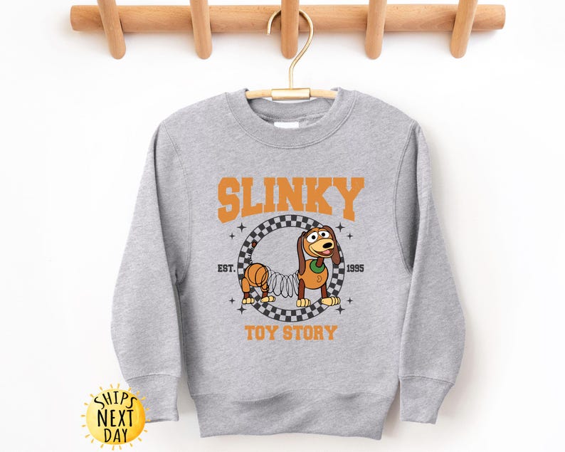 May include: Gray sweatshirt with a graphic of Slinky Dog from Toy Story. The graphic features Slinky Dog in a checkered circle with the text "Slinky" above and "Toy Story" below.