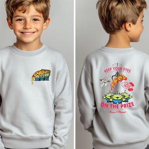 May include: Light gray sweatshirt featuring a colorful graphic of a pizza slice with three green aliens on the front. The back of the sweatshirt has the text "KEEP YOUR EYES ON THE PRIZE" with a claw machine and more aliens.