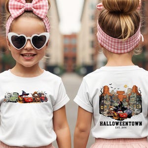 K&ouml;nnte beinhalten: Wei&szlig;es T-Shirt mit einem Halloween-Motiv von Cartoon-Autos. Die Vorderseite zeigt verkleidete Autos. Die R&uuml;ckseite zeigt eine detaillierte Stadtillustration mit dem Text "Halloweentown Est. 2006".