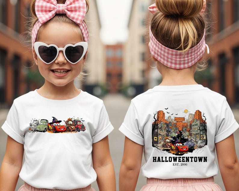 K&ouml;nnte beinhalten: Wei&szlig;es T-Shirt mit einem Halloween-Motiv von Cartoon-Autos. Die Vorderseite zeigt verkleidete Autos. Die R&uuml;ckseite zeigt eine detaillierte Stadtillustration mit dem Text "Halloweentown Est. 2006".