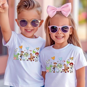 May include: Two children wearing white t-shirts with a colorful Toy Story graphic. The graphic features cartoon characters like Woody, Buzz Lightyear, and others. Both children are wearing sunglasses and smiling.