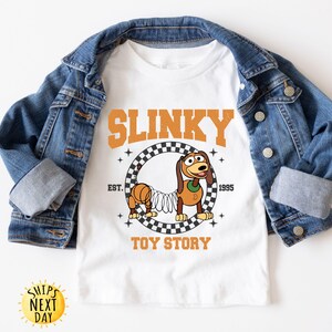 May include: White t-shirt with a graphic of Slinky Dog from Toy Story. The graphic features Slinky Dog on a checkered background with the text "Slinky" above and "Toy Story" below.