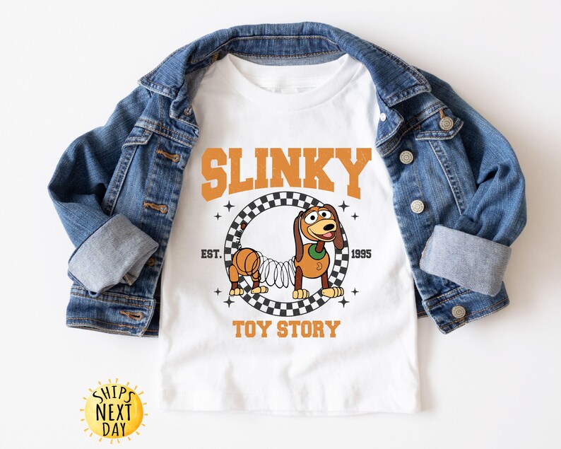 May include: White t-shirt with a graphic of Slinky Dog from Toy Story. The graphic features Slinky Dog on a checkered background with the text "Slinky" above and "Toy Story" below.