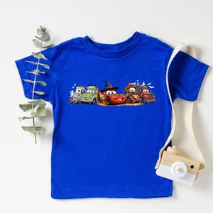 K&ouml;nnte beinhalten: Ein k&ouml;nigsblaues T-Shirt mit einem bunten Design von animierten Autos, darunter ein rotes Auto mit Hexenhut. Das Design ist auf dem Shirt zentriert, mit den Autos in einer Reihe angeordnet. Das Shirt ist aus weichem Material.
