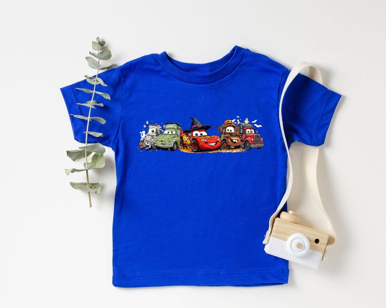 K&ouml;nnte beinhalten: Ein k&ouml;nigsblaues T-Shirt mit einem bunten Design von animierten Autos, darunter ein rotes Auto mit Hexenhut. Das Design ist auf dem Shirt zentriert, mit den Autos in einer Reihe angeordnet. Das Shirt ist aus weichem Material.