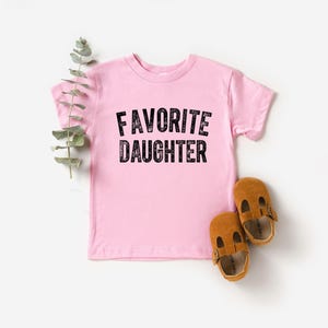 May include: A pale pink toddler t-shirt with the text "FAVORITE DAUGHTER" in black. Two brown leather baby shoes are next to the shirt. A sprig of green leaves is placed near the shirt.