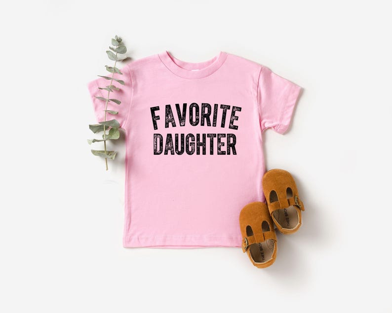 May include: A pale pink toddler t-shirt with the text "FAVORITE DAUGHTER" in black. Two brown leather baby shoes are next to the shirt. A sprig of green leaves is placed near the shirt.