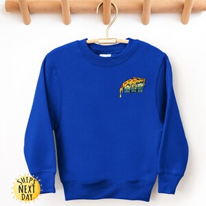 May include: Royal blue sweatshirt with a crew neck. The sweatshirt features a small, colorful graphic of a pizza slice with a cartoon face. The sweatshirt is hanging on a wooden hanger.