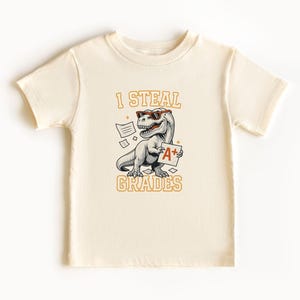 May include: Cream-colored t-shirt featuring a cartoon dinosaur wearing sunglasses and holding an A+ paper. The text "I STEAL GRADES" is above the dinosaur, with "GRADES" below. The design is in shades of gray, orange, and black.