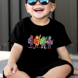 May include: Black t-shirt featuring a colorful cartoon illustration of Spider-Man, Iron Man, Hulk, and Captain America, each holding a pencil and wearing a backpack. The design is centered on the front of the shirt.