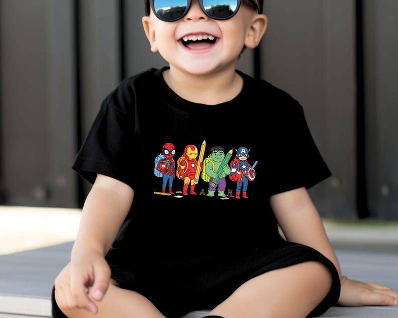 May include: Black t-shirt featuring a colorful cartoon illustration of Spider-Man, Iron Man, Hulk, and Captain America, each holding a pencil and wearing a backpack. The design is centered on the front of the shirt.