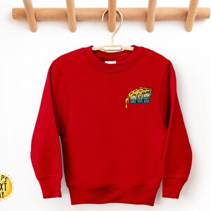 May include: A bright red sweatshirt with a small, colorful pizza slice graphic on the upper left chest. The pizza slice has melted cheese and a cartoon design. The sweatshirt is hanging on a wooden hanger.