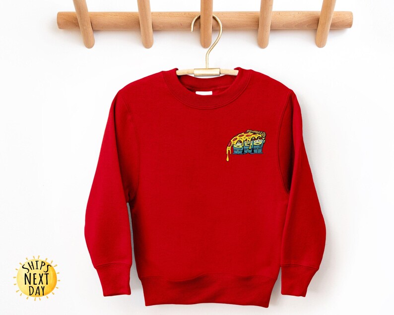 May include: A bright red sweatshirt with a small, colorful pizza slice graphic on the upper left chest. The pizza slice has melted cheese and a cartoon design. The sweatshirt is hanging on a wooden hanger.