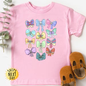May include: Pink t-shirt with a colorful bow design. The bows are in various colors and patterns. The shirt has a crew neck and short sleeves.