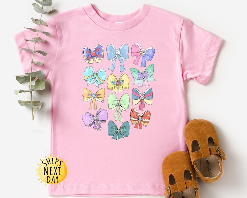 May include: Pink t-shirt with a colorful bow design. The bows are in various colors and patterns. The shirt has a crew neck and short sleeves.