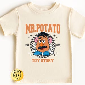 May include: A cream colored t-shirt with a cartoon image of Mr. Potato Head from Toy Story. The text "MR. POTATO" is in orange letters above the image and "TOY STORY" is in orange letters below the image. The text "EST. 1995" is in black letters to the right of the image.