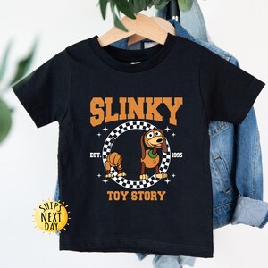 May include: Black t-shirt with a cartoon image of Slinky Dog from Toy Story. The shirt has the text "Slinky" in a white outline with a black fill, "Est. 1995" in white text, and "Toy Story" in white text. The image of Slinky Dog is in a checkered circle.
