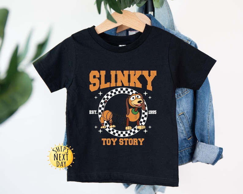 May include: Black t-shirt with a cartoon image of Slinky Dog from Toy Story. The shirt has the text "Slinky" in a white outline with a black fill, "Est. 1995" in white text, and "Toy Story" in white text. The image of Slinky Dog is in a checkered circle.