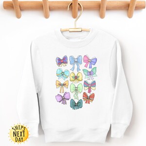 May include: White sweatshirt with a colorful bow design. The bows are in various colors, including pink, blue, yellow, and green. The sweatshirt has a crew neck and long sleeves.
