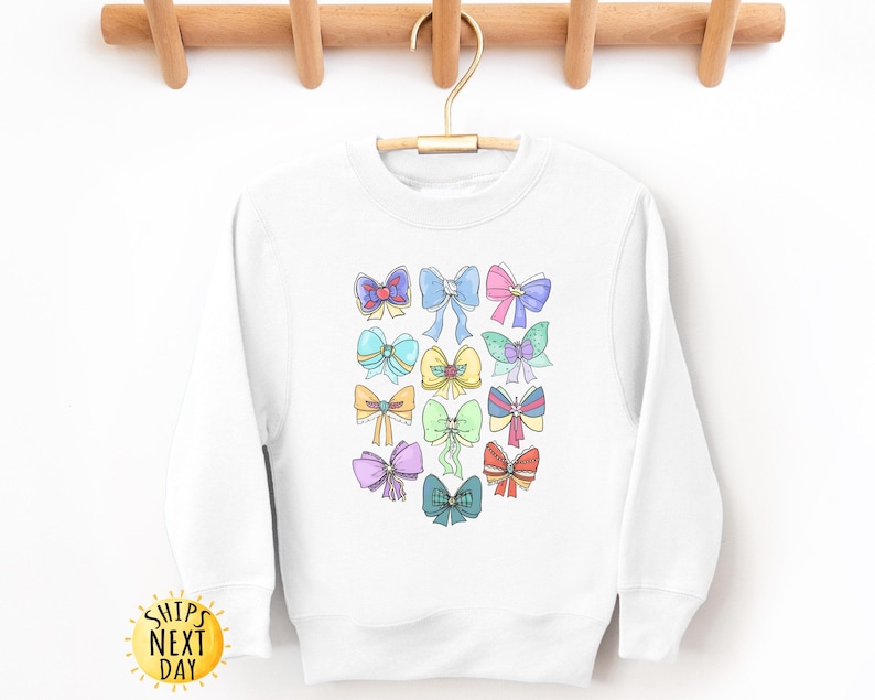 May include: White sweatshirt with a colorful bow design. The bows are in various colors, including pink, blue, yellow, and green. The sweatshirt has a crew neck and long sleeves.