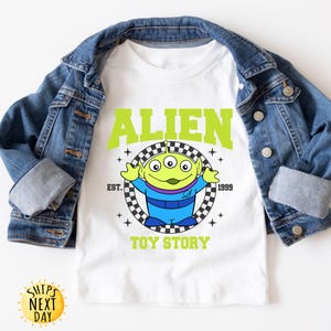 Puede incluir: Camiseta blanca con un personaje extraterrestre verde de Toy Story. La camiseta tiene el texto "Alien" en letras verdes con un patr&oacute;n a cuadros y el texto "Toy Story" debajo.