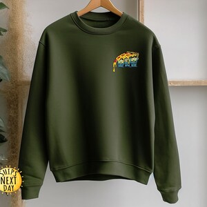 May include: Olive green sweatshirt with a crew neck and long sleeves. A colorful graphic of a pizza slice with faces is on the upper left chest. The sweatshirt is hanging on a wooden hanger.