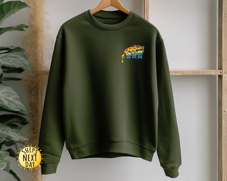 May include: Olive green sweatshirt with a crew neck and long sleeves. A colorful graphic of a pizza slice with faces is on the upper left chest. The sweatshirt is hanging on a wooden hanger.