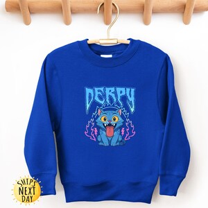 May include: Royal blue sweatshirt with a cartoon cat graphic and the word "DERPY" in blue. The cat has a pink tongue and is surrounded by pink and blue flames. The sweatshirt is displayed on a wooden hanger.
