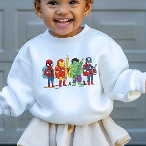 May include: White crewneck sweatshirt featuring a colorful graphic of cartoon superheroes, including Spider-Man, Iron Man, Hulk, and Captain America, each holding school supplies. The design is centered on the front of the sweatshirt.