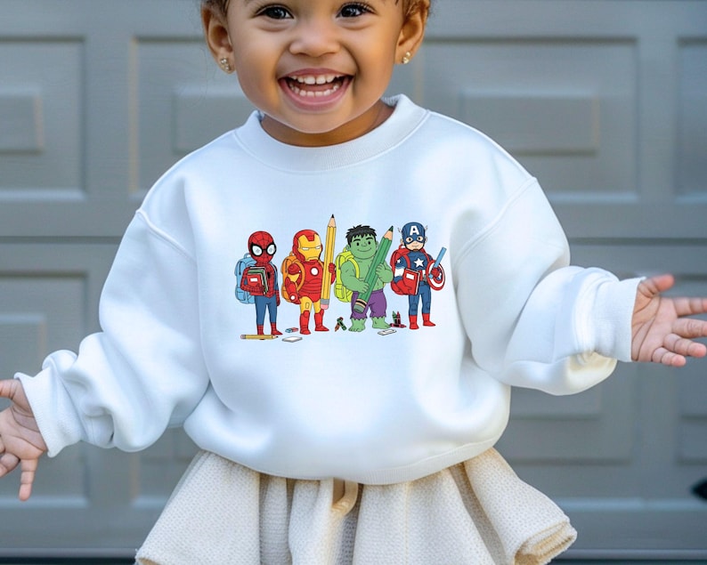 May include: White crewneck sweatshirt featuring a colorful graphic of cartoon superheroes, including Spider-Man, Iron Man, Hulk, and Captain America, each holding school supplies. The design is centered on the front of the sweatshirt.