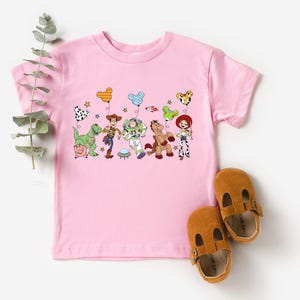 May include: A light pink toddler t-shirt featuring a colorful graphic of Toy Story characters holding balloons. The design includes Woody, Buzz Lightyear, Jessie, and other characters. Brown leather baby shoes are next to the shirt.