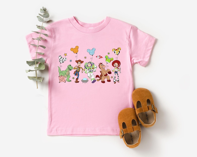 May include: A light pink toddler t-shirt featuring a colorful graphic of Toy Story characters holding balloons. The design includes Woody, Buzz Lightyear, Jessie, and other characters. Brown leather baby shoes are next to the shirt.